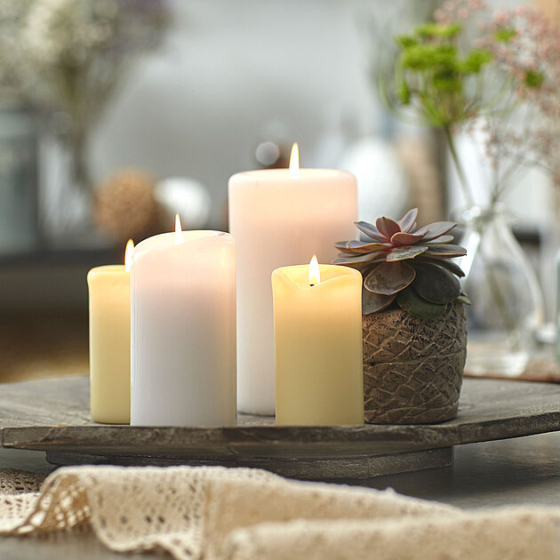Auf einem runden Holzteller stehen vier brennende Bolsius Stumpenkerzen in Weiß- und Cremetönen, begleitet von einem kleinen Sukkulenten-Topf. Im Vordergrund liegt eine gehäkelte Tischborte, im Hintergrund sind unscharf Blumen in Glasvasen und Gebäck auf einer Etagere zu erkennen. Die Szene wirkt hell, freundlich und dekorativ.