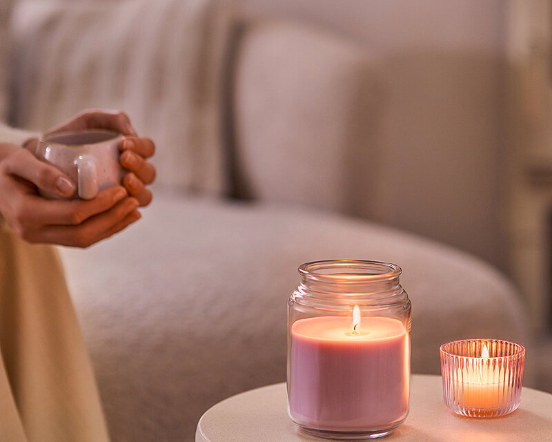 Gemütliche Wohnszene mit einer brennenden Bolsius True Scents Duftkerze im Bonbonglas und einem Teelicht auf einem Beistelltisch neben einer Person mit einer Tasse.