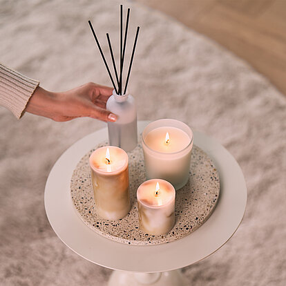 Stilvolle Wohnszene mit der Marble-Kollektion von Bolsius in unterschiedlichen Größen und einem Raumduft-Diffuser auf einem runden Beistelltisch. Eine Hand stellt den Diffuser auf den Tisch. Warmes Kerzenlicht und dezente Naturtöne schaffen eine ruhige, elegante Wohlfühlatmosphäre