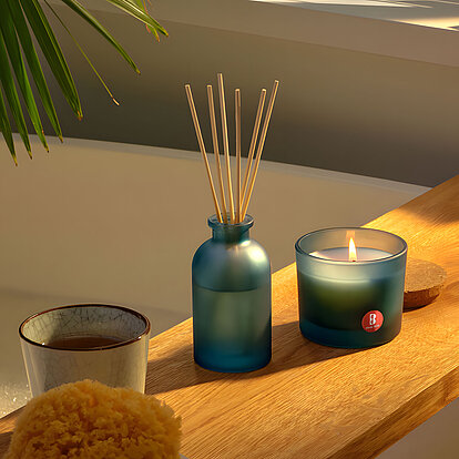 Ein entspannendes Badezimmer-Setting im sanften Tageslicht: Ein zart bläuliches Glasgefäß mit Duftstäbchen steht mittig auf einem rustikalen Holzboard über der schaumigen Badewanne. Daneben flackert eine Kerze im passenden Blauton. Das Arrangement umgibt Moos-Schwamm, Teebecher und seifige Accessoires, eingerahmt von Palmenblatt-Schatten und Jalousien-Licht – ganz auf die beruhigende Wirkung des Raumdufts von Bolsius ausgerichtet.