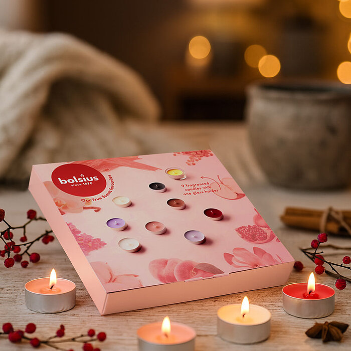 Eine rosafarbene Geschenkbox mit Duft-Teelichtern von Bolsius liegt auf einem Holztisch. Darum brennen mehrere Teelichter, dekoriert mit roten Beeren, Zimtstangen und Sternanis. Im Hintergrund sorgen Lichter für gemütliche Stimmung.