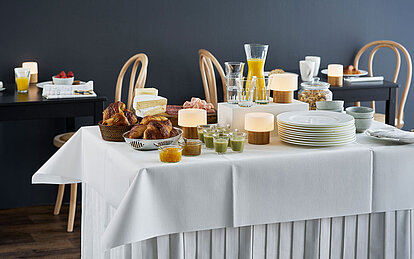 Auf einem reichlich dekorierten Tisch mit einer weißen Tischdecke sind unterschiedliche Nahrungsmittel wie Brot, Joghurt, Brötchen und einer Karaffe mit Orangensaft. Daneben steht ein großer Stapel mit weißen Tellern. Im Hintergrund sind weitere Regale mit einer Auswahl an Frühstücksnahrungsmitteln.