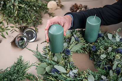 Ein großer Kranz aus vielen grünen zweigen, Ästen und Blättern auf dem zwei grüße große Stumpenkerzen stehen. Man sieht wie eine menschliche Hand eine der Kerzen gerade auf dem Kranz anbringt. Um den Kranz herum liegen mehrere Äste und zweige verteilt.