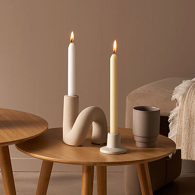 Stilvoll dekorierte Spitzkerzen und Tafelkerzen brennen in modernen Haltern auf zwei Holztischen. Die Kerzen in sanften Naturtönen schaffen eine ruhige und gemütliche Atmosphäre. Kombiniert mit Deko-Elementen wie getrockneten Mohnkapseln, Keramikvasen und weichen Textilien, passen sie perfekt in ein minimalistisches Wohnambiente. Ideal für stilvolle Tischdekoration oder entspannte Abende.