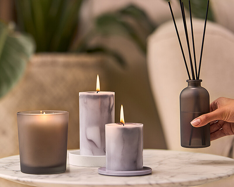 Bolsius Marble Kollektion als stilvolle Wohnraumdekoration mit Duftkerze im Glas, Marmoreffekt-Stumpenkerzen und Reed Diffuser. Elegante Dekoration auf einem Marmortisch für eine gemütliche Atmosphäre und modernes Wohnambiente.