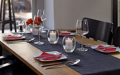 bolsius-kerzen-gastronomie-impression-2023-1 Die Kerzen Gastronomie setzt auf eine stilvolle Tischdekoration in einem Restaurant, die mit weißen Tellern und roten Servietten anspruchsvoll gestaltet ist. Auf dem Tisch steht eine weiße Vase mit roten Blumen und leere Weingläser, begleitet von zwei brennenden Relight Kerzen in einem ovalen Glashalter, die eine gemütliche und einladende Stimmung verbreiten.