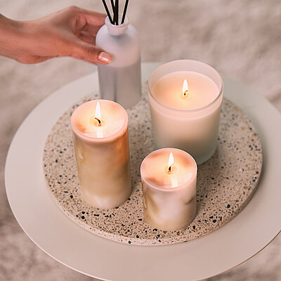 Wohnszene mit drei stilvollen Marble-Kollektion Stumpenkerzen von Bolsius in unterschiedlichen Größen und einem Raumduft-Diffuser auf einem runden Beistelltisch. Eine Hand stellt den Diffuser auf den Tisch. Warmes Kerzenlicht und dezente Naturtöne schaffen eine ruhige, elegante Wohlfühlatmosphäre.