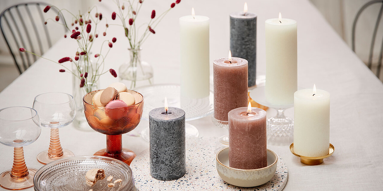 Auf einem elegant gedeckten Tisch stehen mehrere Bolsius Professional Rustik-Stumpenkerzen in natürlichen Farbtönen wie Creme, Grau und Rosé. Die grobe, strukturierte Oberfläche der Kerzen unterstreicht den handgefertigten Charakter und sorgt für ein stilvolles, authentisches Ambiente. Umgeben von Glasgeschirr, Macarons und zarten Trockenblumen, erzeugen die brennenden Kerzen eine warme, einladende Atmosphäre – ideal für stilvolle Gastronomie, Events oder festliche Tischdekorationen.