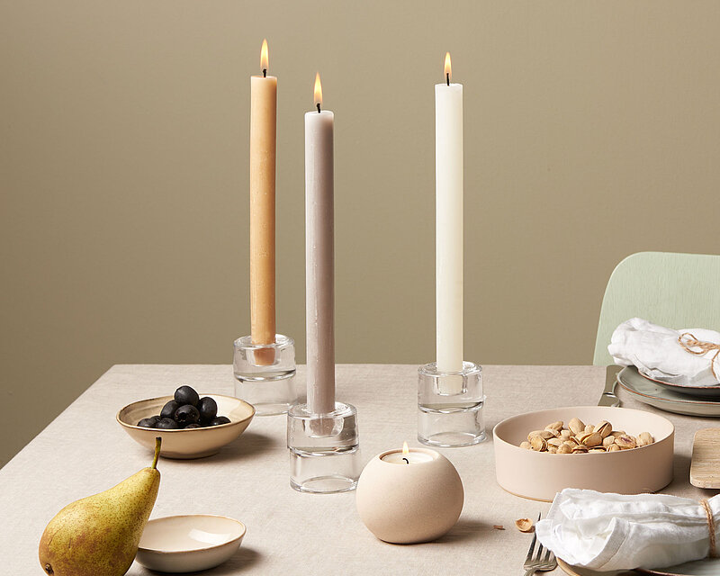 Drei brennende Bolsius Rustik Haushaltskerzen bzw. Stabkerzen in sanften Naturfarben stehen in schlichten Glas-Kerzenhaltern auf einem gedeckten Esstisch. Die Kombination aus edler Tischdecke, schlichtem Geschirr und natürlichen Lebensmitteln wie Birne, Nüssen und Trauben schafft eine ruhige und moderne Atmosphäre. 