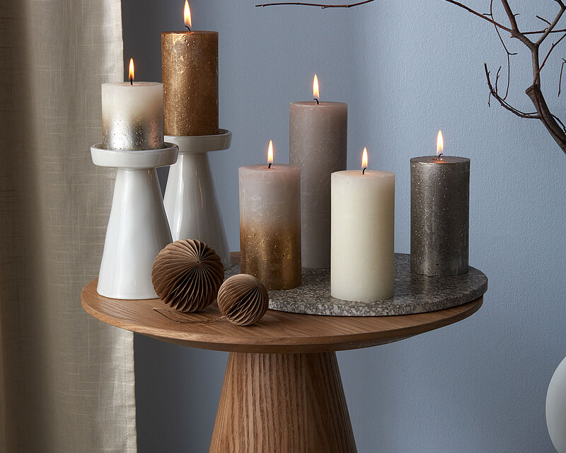 Mehrere elegante Bolsius metallic Rustik-Stumpenkerzen in Gold, Silber, Grau und Creme stehen brennend auf einem modernen Beistelltisch. Die Kombination aus glänzenden und matten Oberflächen sorgt für einen edlen Look. Die Kerzen passen perfekt zur Winter- und Weihnachtsdekoration und schaffen eine warme, stilvolle Atmosphäre im Wohnraum. 