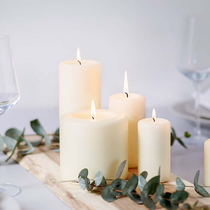 Mehrere cremefarbene Stumpenkerzen brennen stimmungsvoll auf einem Holztablett und sorgen für ein warmes Licht. Die Kerzen sind umgeben von Eukalyptuszweigen und stehen auf einem elegant gedeckten Tisch mit Weingläsern und Tellern.