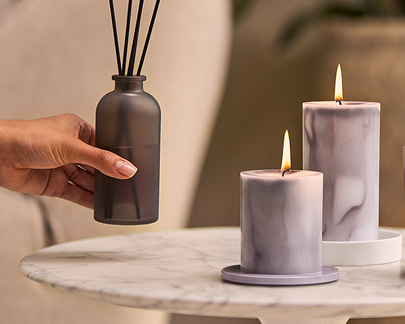 Elegante Wohnszene mit mattschwarzem Raumduft-Diffuser aus der Bolsius Marble Kollektion und zwei marmorierten Stumpenkerzen auf einem runden Marmortisch. Eine Hand platziert den Diffuser und schafft eine stilvolle, ruhige Wohlfühlatmosphäre im modernen Zuhause.
