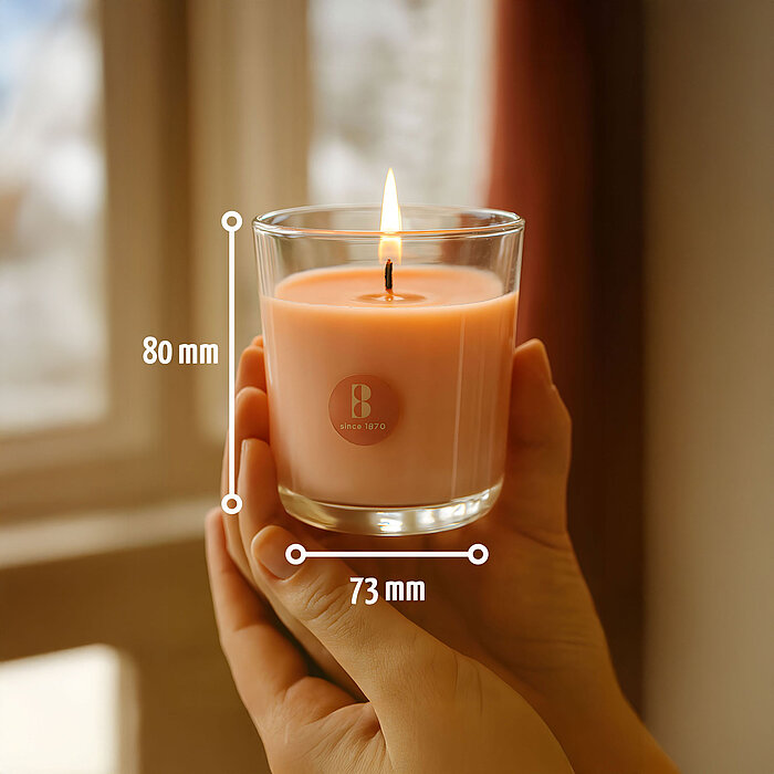 Zwei zarte Hände halten eine brennende Duftkerze im Glas von Bolsius. Auf dem Bild wird die Größe von 80/73mm (H/DM) dargestellt.