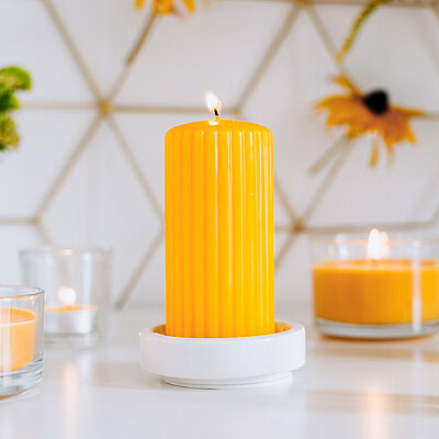 Im Fokus eine leuchtend gelbe Stumpenkerze von Bolsius, platziert auf einem eleganten Keramikteller. Darum angeordnet stehen weitere Duftkerzen im Glas‑ und Teelichtformat, die gemeinsam ein angenehmes Duftambiente schaffen. Dezente Blumen in kleinen Vasen und ein dezent grafisch gestalteter Hintergrund ergänzen das stilvolle Gesamtbild – ideal für harmonische Wohlfühlmomente mit aromatischen Duft-Stumpenkerzen von Bolsius.