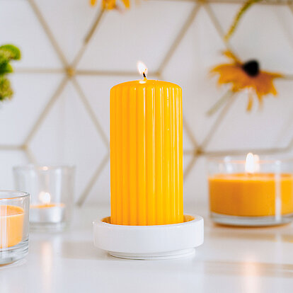 Im Fokus eine leuchtend gelbe Stumpenkerze von Bolsius, platziert auf einem eleganten Keramikteller. Darum angeordnet stehen weitere Duftkerzen im Glas‑ und Teelichtformat, die gemeinsam ein angenehmes Duftambiente schaffen. Dezente Blumen in kleinen Vasen und ein dezent grafisch gestalteter Hintergrund ergänzen das stilvolle Gesamtbild – ideal für harmonische Wohlfühlmomente mit aromatischen Duft-Stumpenkerzen von Bolsius.